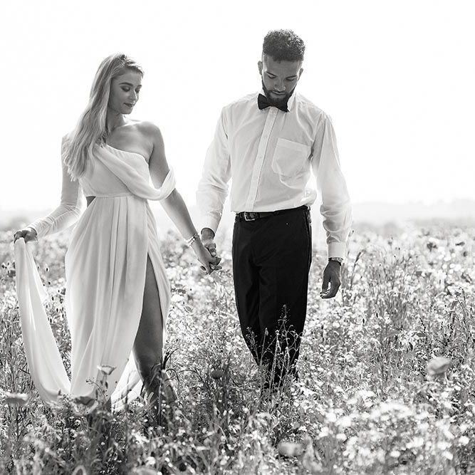 Black and white portrait of the bride and groom holding hands in a field. The bride wears a wrap wedding dress with front split and one shoulder detail 