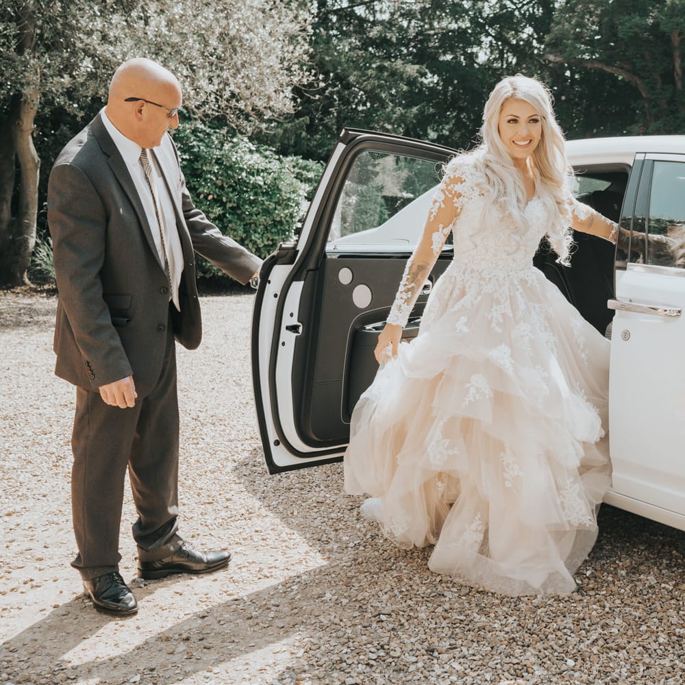 A bride in a tiered tulle wedding dress steps out of a white wedding car. 