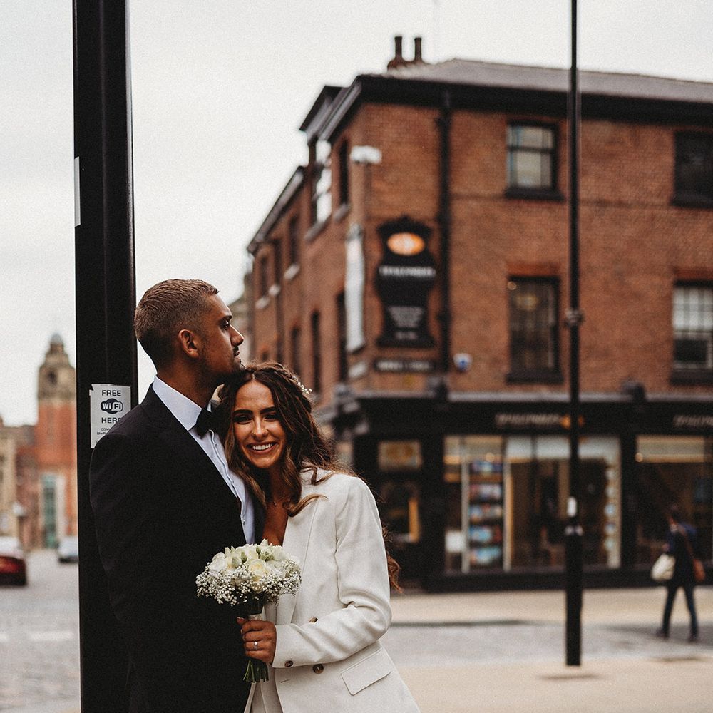 Bride in a wide leg trouser suit holding a white flower bouquet embracing her groom in a black tuxedo 