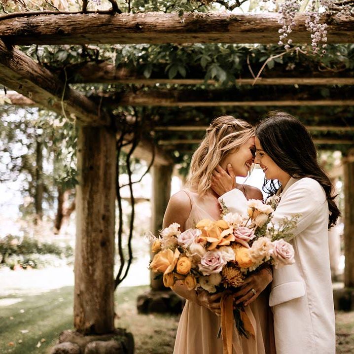 Bride in a white suit kissing her bride in a blush pink wedding dress under a pergola 
