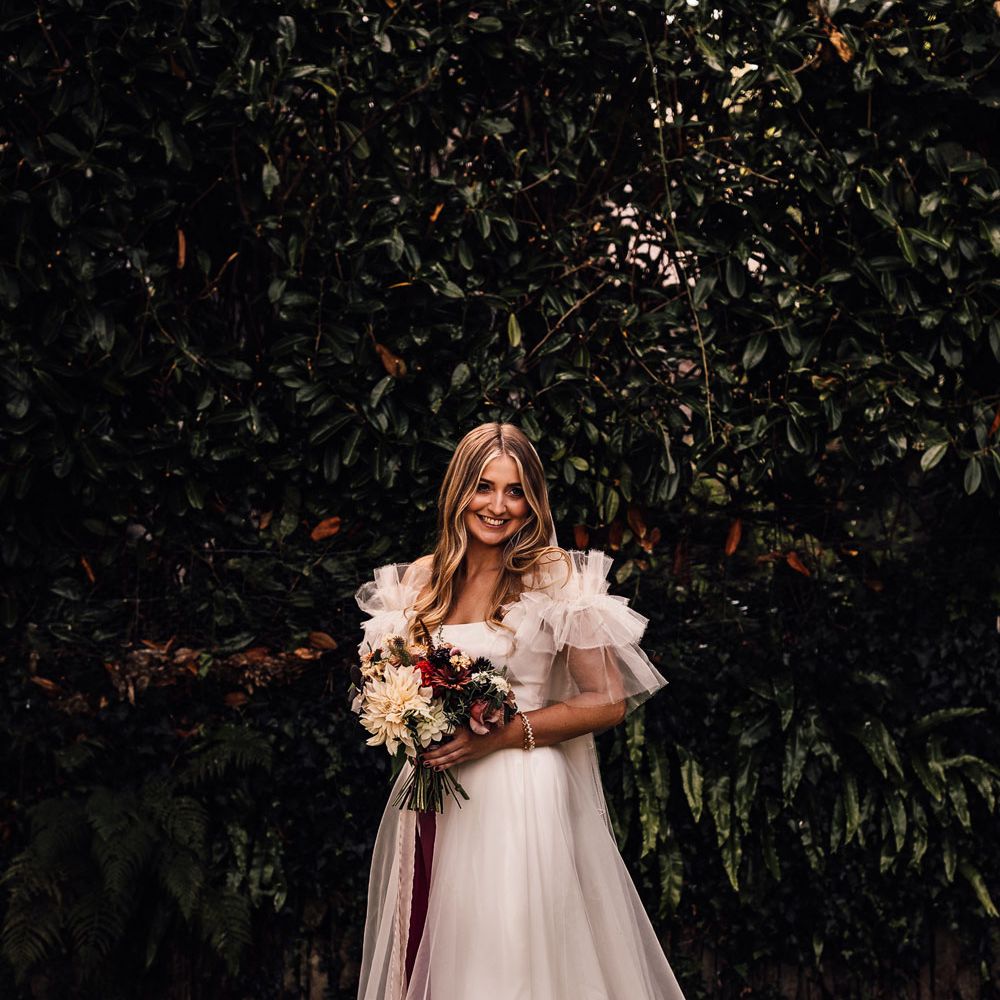 Bride in white Halfpenny London Mayfair dress stands outside holding mixed bridal bouquet with burgundy ribbon before church wedding ceremony in Cornwall