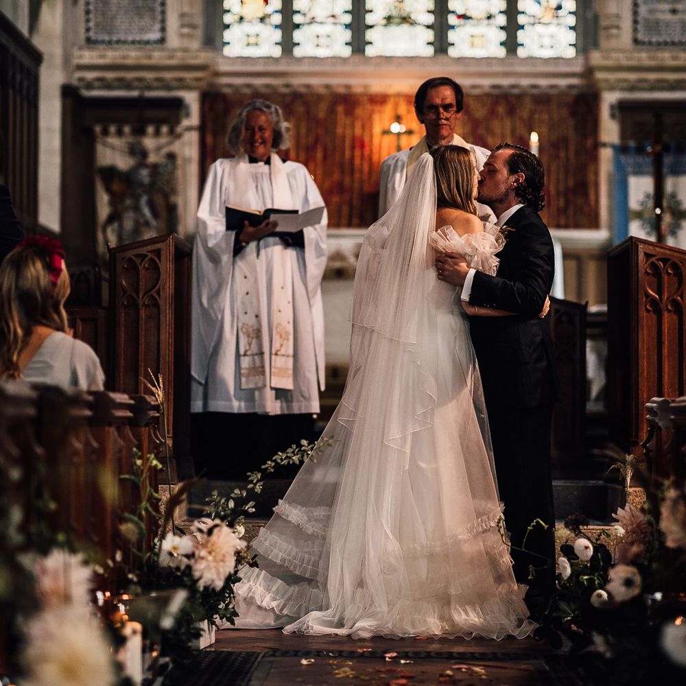 Bride in white Halfpenny London Mayfair dress and veil kisses groom in three piece suit at the altar during church ceremony in Cornwall 