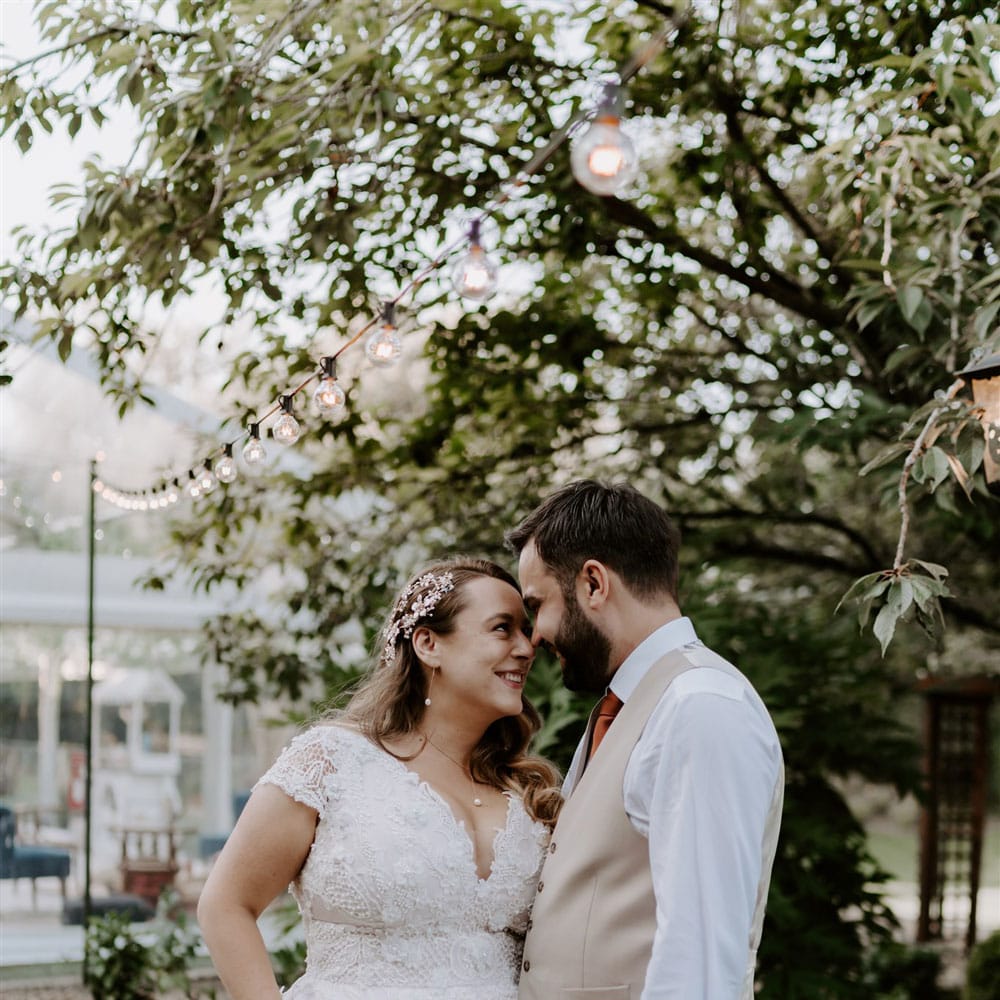 Bride in homemade lace wedding dress with pearl floral hair pin smiles at groom in beige suits trousers and waistcoat as they stand in garden after wedding ceremony