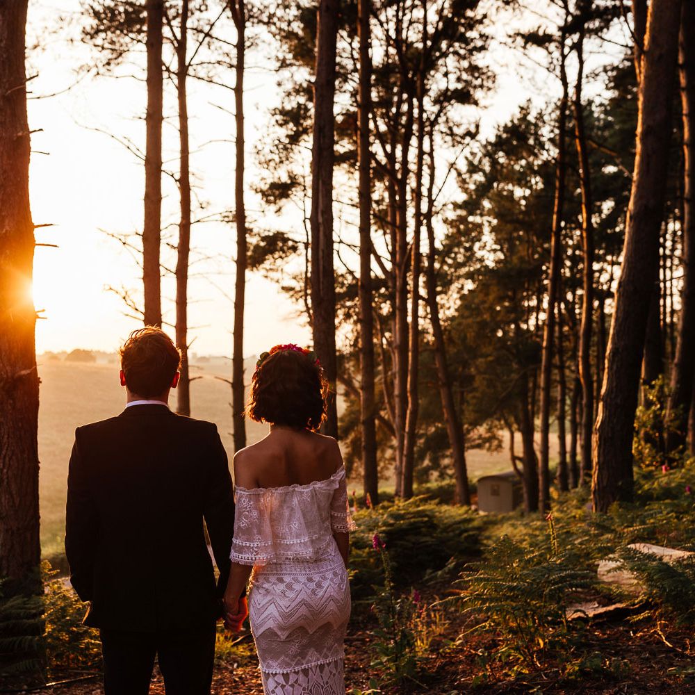 Bride in off the shoulder Grace Loves Lace wedding dress and flower crown stands looking towards the sun holding hands with groom in black suit during golden hour