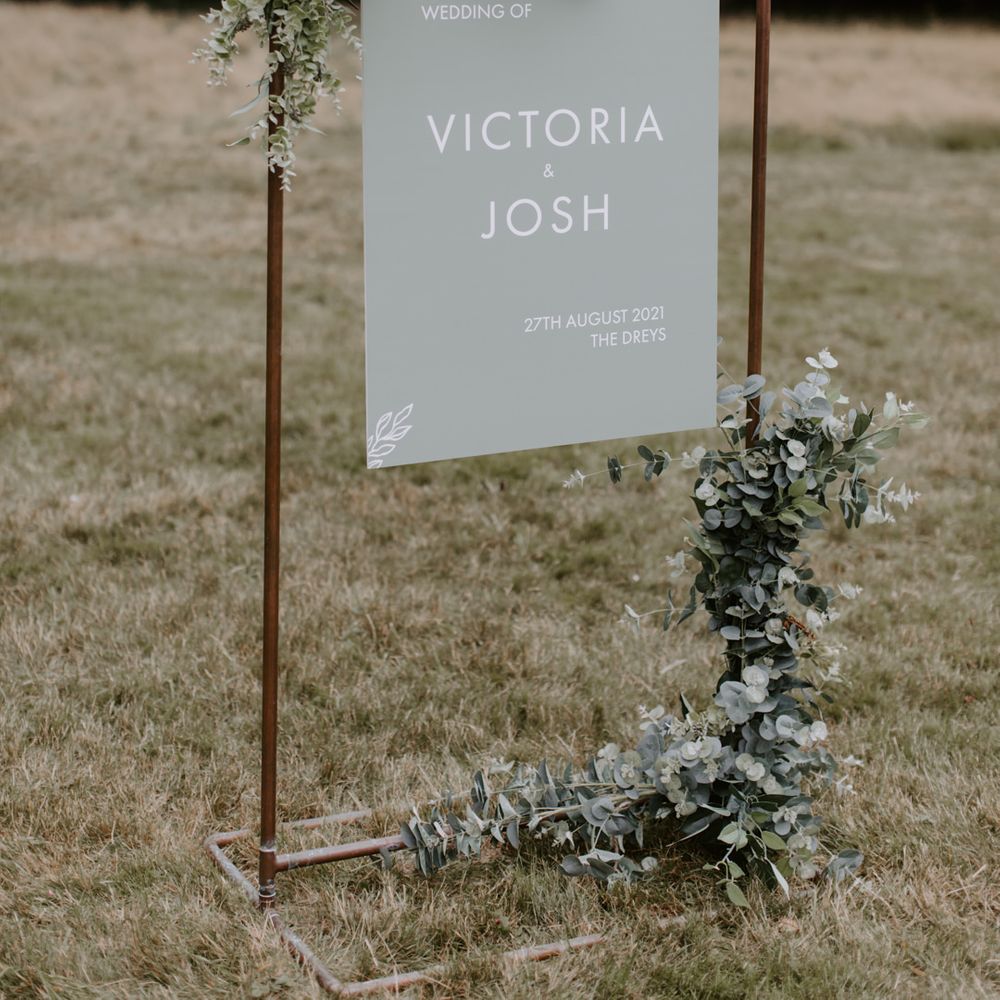 Welcome sign complete in sage green held by brass stand and surrounded by foliage