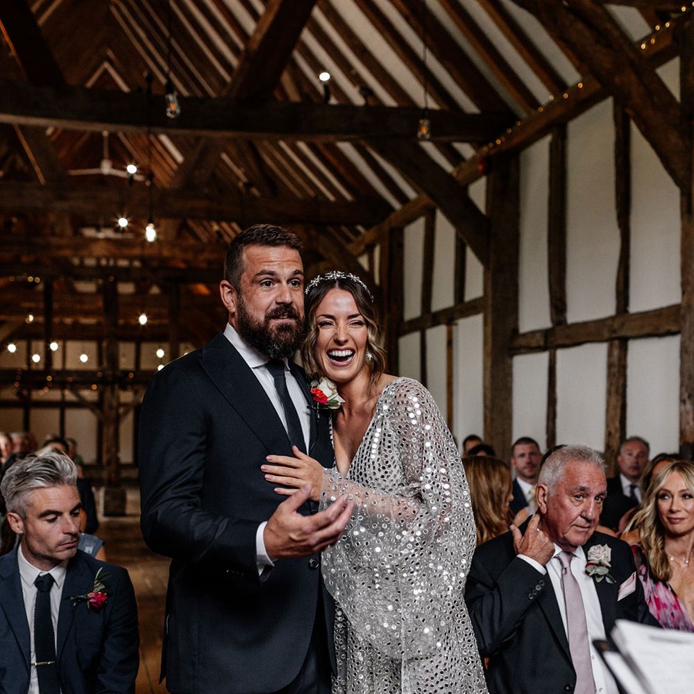 Bride in a silver sequin wedding dress laughing during the barn wedding ceremony with her groom in a navy suit 