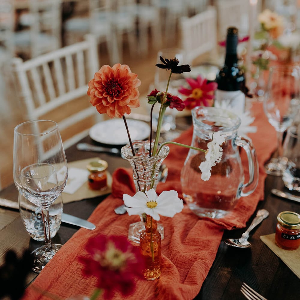 Orange table runner lines wooden table alongside brightly coloured flowers 