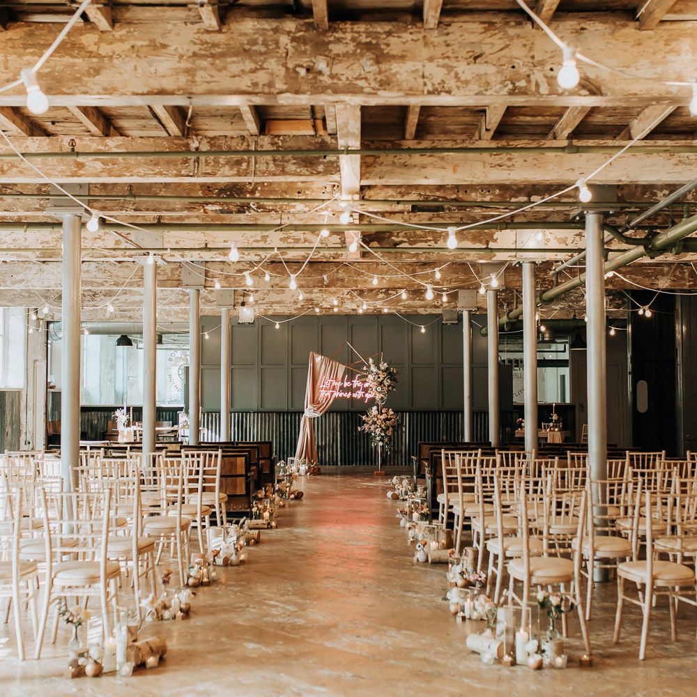 Industrial wedding ceremony room with pillars, wooden chairs, pastel florals and pink neon wedding sign