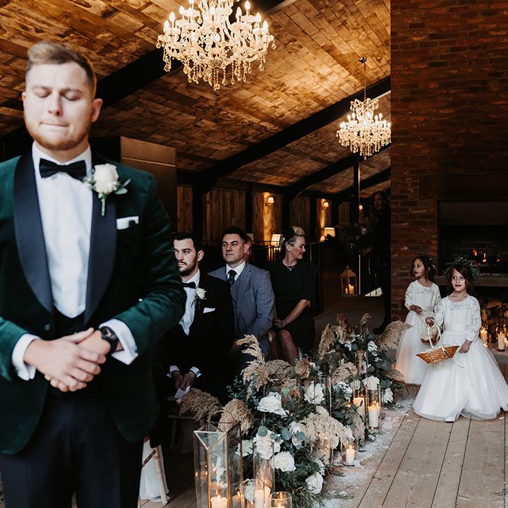 Flower girls in lace white tulle princess dresses walk down the aisle whilst the groom nervously waits
