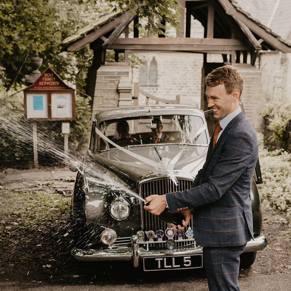 Groomsman in blue and brown checkered suit and orange tie pops some champagne with bride and groom in grey wedding car in the background 
