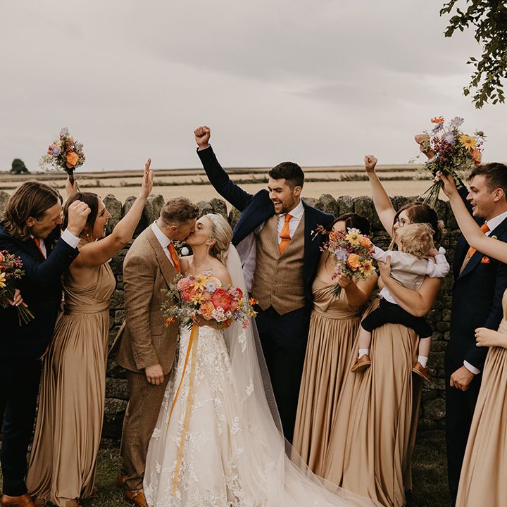 Bride and groom share a kiss as the wedding party cheer them on with bridesmaids in gold dresses and colourful flowers 