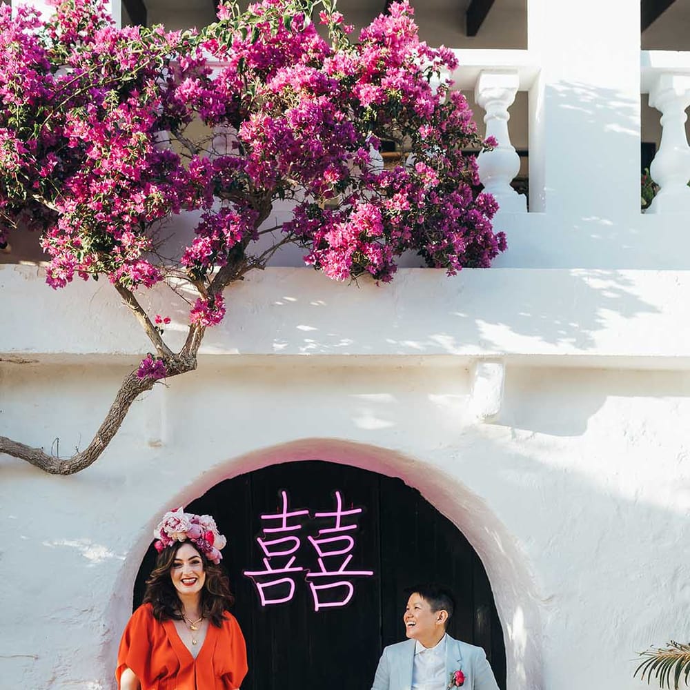 Newlyweds stand in front of pink LED light with Chinese symbols as they wear red gown and a pale blue suit 