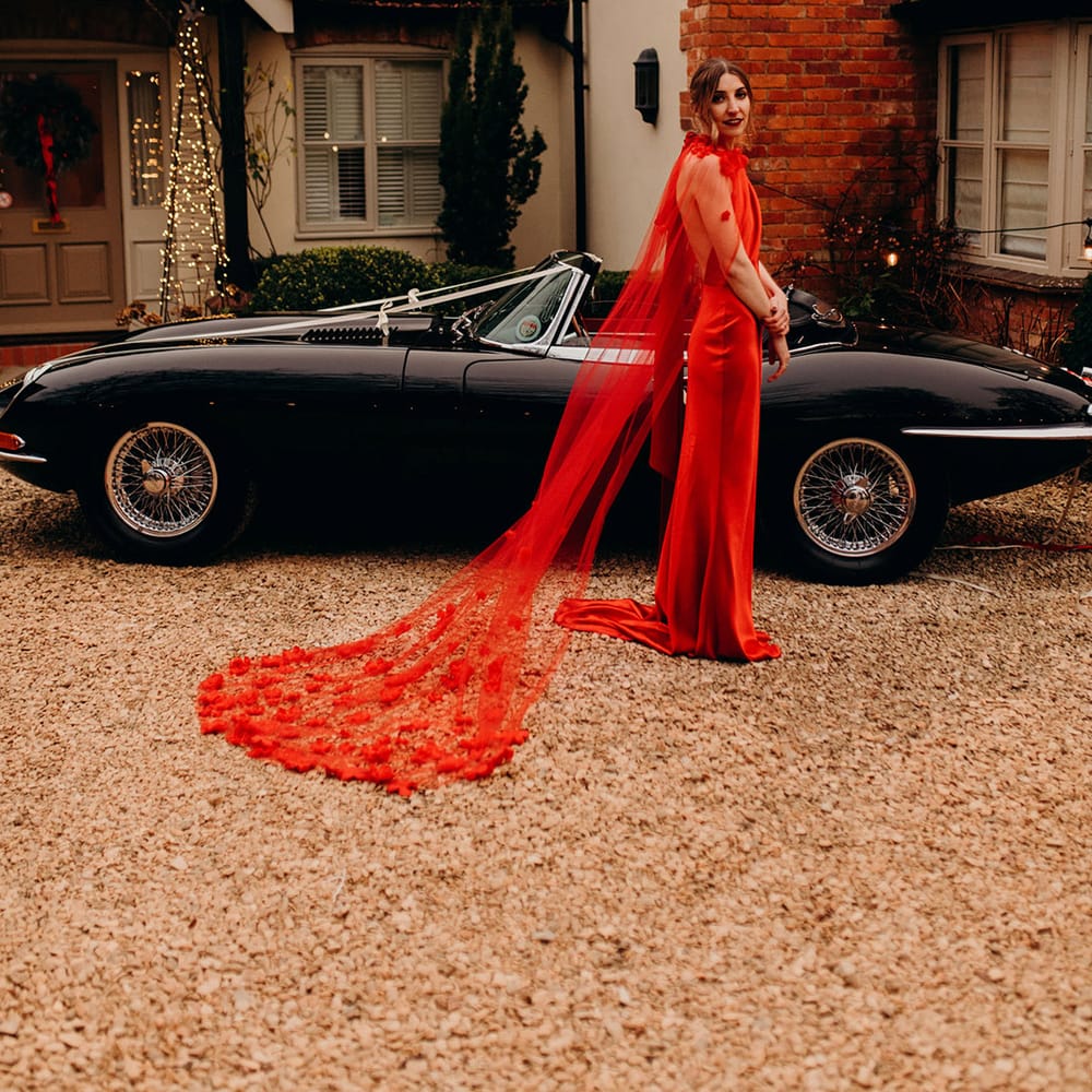 Bride in satin red wedding dress with cape and 3d appliqué roses standing by classic convertible wedding car