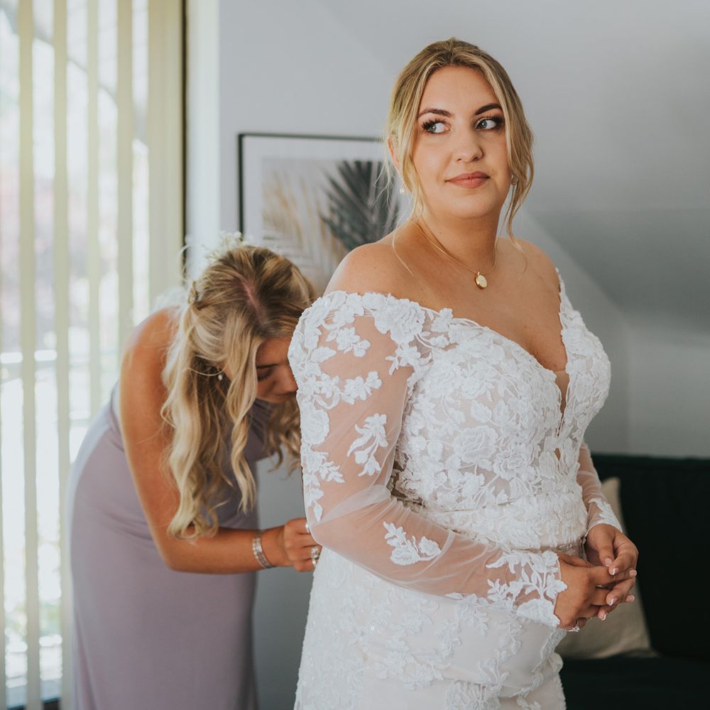 Bridesmaid in pink dress helps bride do up the back of her lace wedding dress