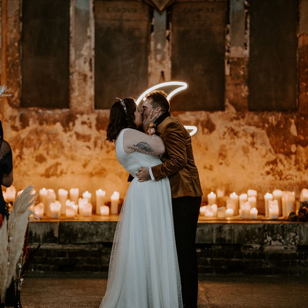 Bride and Groom share a kiss at unique wedding venue. Their wedding lighting is created by candles and a moon neon sign