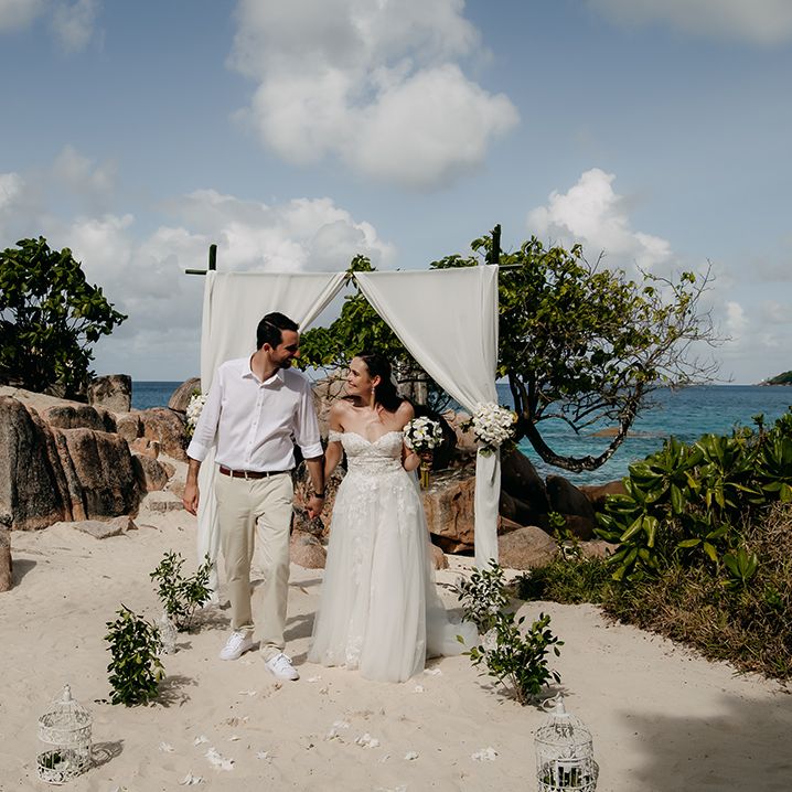 Groom in crisp white shirt, light coloured chinos, tan leather belt and white trainers holding hands with bride  in off the shoulder lace wedding dress with sweetheart neckline and lace floral details and cathedral length veil holding white tropical bouquet 