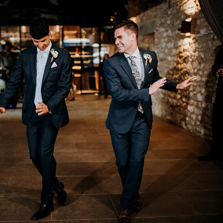 Grooms in blue suits and white floral buttonholes dance during wedding reception in barn