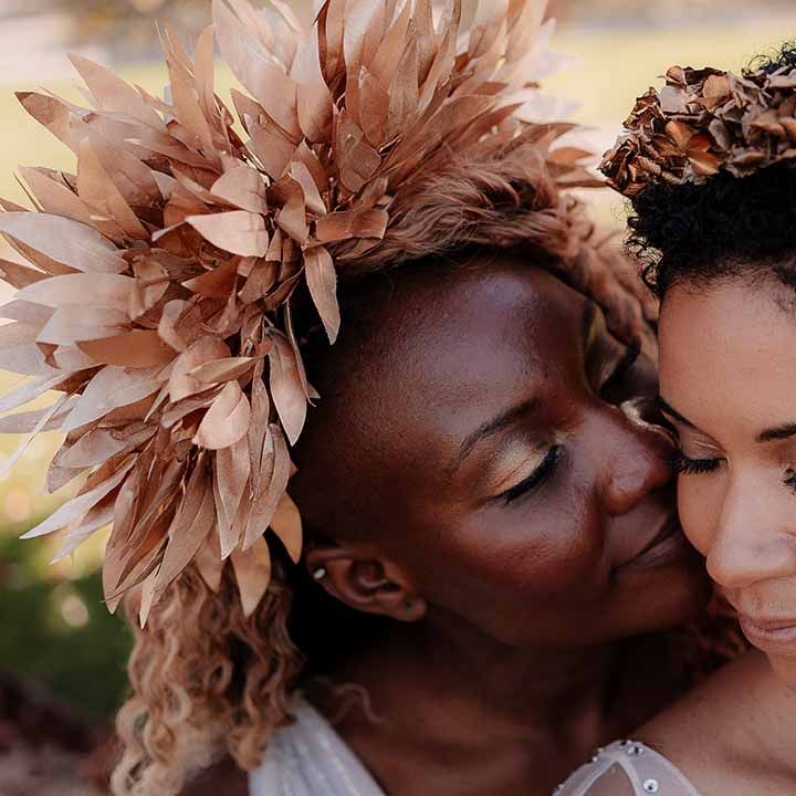 Bride in large golden leaf bridal crown kissing bride in golden floral bridal headpiece on the cheek