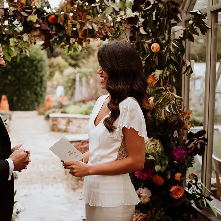 Bride and groom reading their vows at La Manoir Aux Quat Saisons standing by large floral arch with white and pink carnations, garden roses, red strawflower, eucalyptus, baby’s breath and various dried flowers 