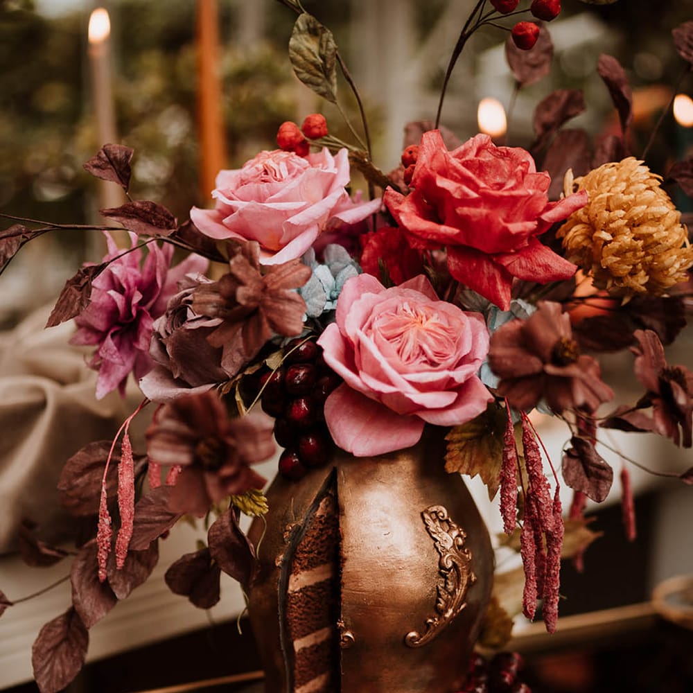 single tier iced wedding cake with intricate leaf details in the style of an aged golden vase filled with handmade wafer paper florals, pink roses, red roses and yellow carnations 