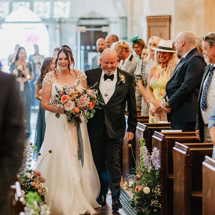 Father of the bride in black tie walks down the aisle with the bride in a flower wedding dress walking down the aisle to the groom at the end 