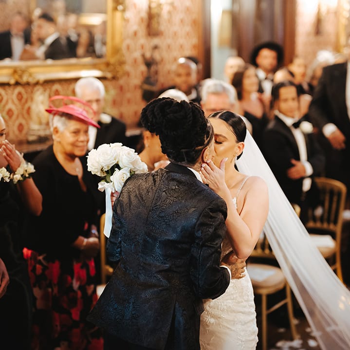 Bride in sleeveless v neck Enzoani wedding dress with tulle finish and church length veil kissing bride in black snakeskin print tuxedo with black bowtie and white rose boutonniere 