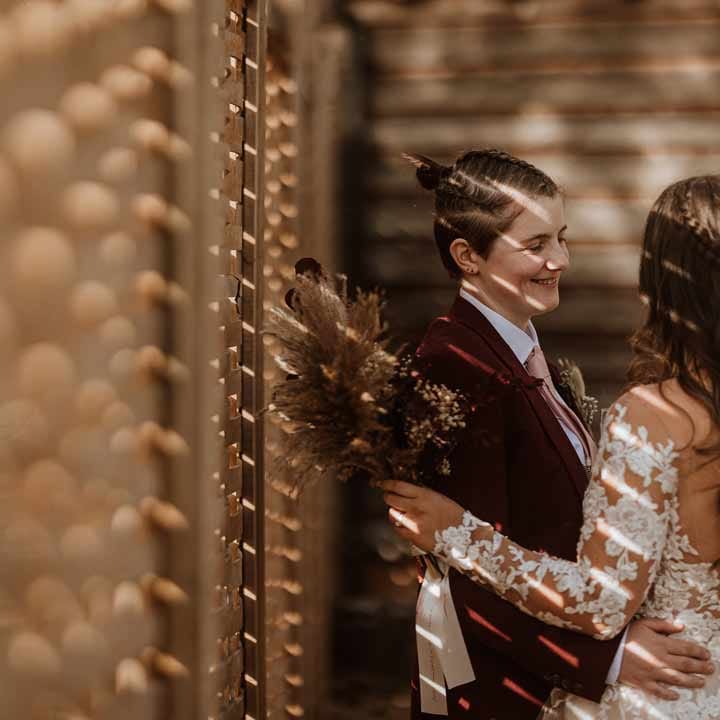 Couples wedding portraits - bride in maroon bridal suit with grey waistcoat, dusky pink tie and autumnal dried flower boutonniere embracing bride in long sleeve lace illusion sleeve wedding dress with overlay holding large autumnal boho dried flower bouquet 