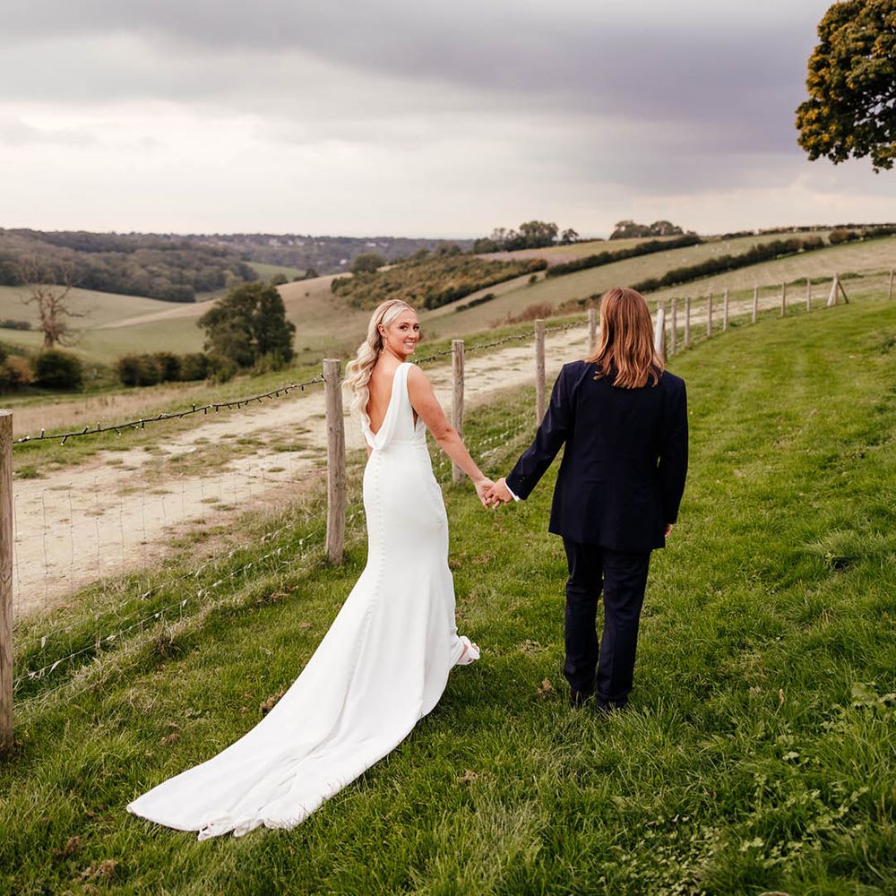 Bride with Hollywood curled blonde hair in draped back wedding dress walking hand in hand with the groom in blue suit 