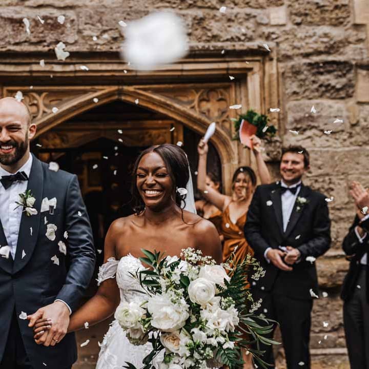 Bride in sweetheart neckline lace wedding dress with detachable off the shoulder sleeves leaving the church with groom in classic black three piece grooms suit with bow tie and white rose and foliage boutonniere doing dried petal confetti exit 