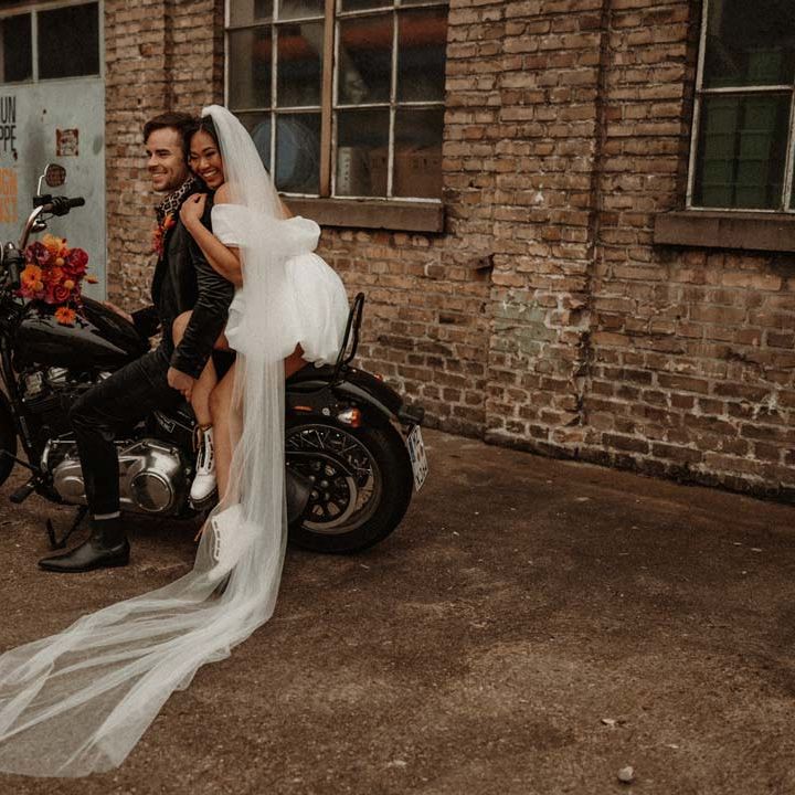 Bride in off shoulder tulle short wedding dress with church length veil and white platform Dr Martens wedding shoes on the back of wedding motorbike with groom in wet look black grooms suit with leopard print shirt and pink and orange boutonniere
