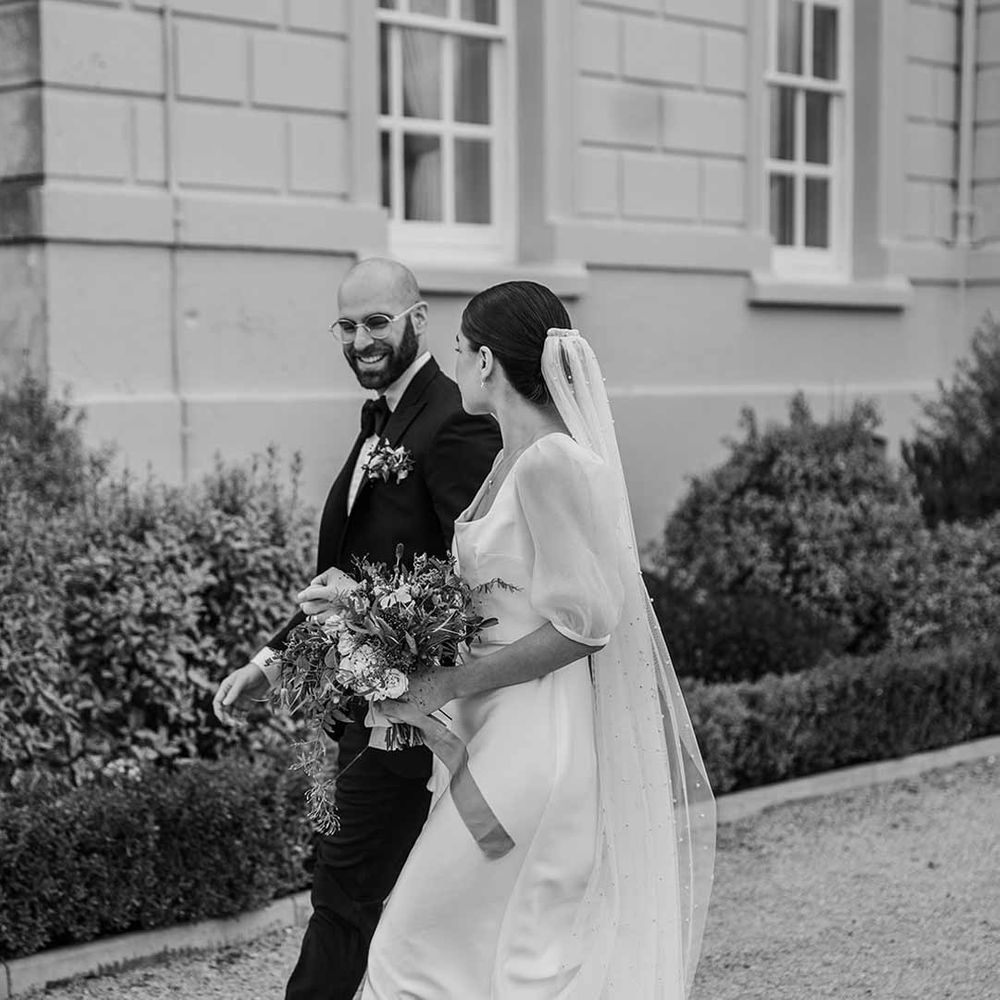 Bride in short sheer puff sleeve wedding dress with square neck and wrist loop with sheer pearl embellished bridal veil holding dried wildflower bouquet walking with groom in classic black tuxedo with bowtie and wildflower boutonniere at Buxted Park