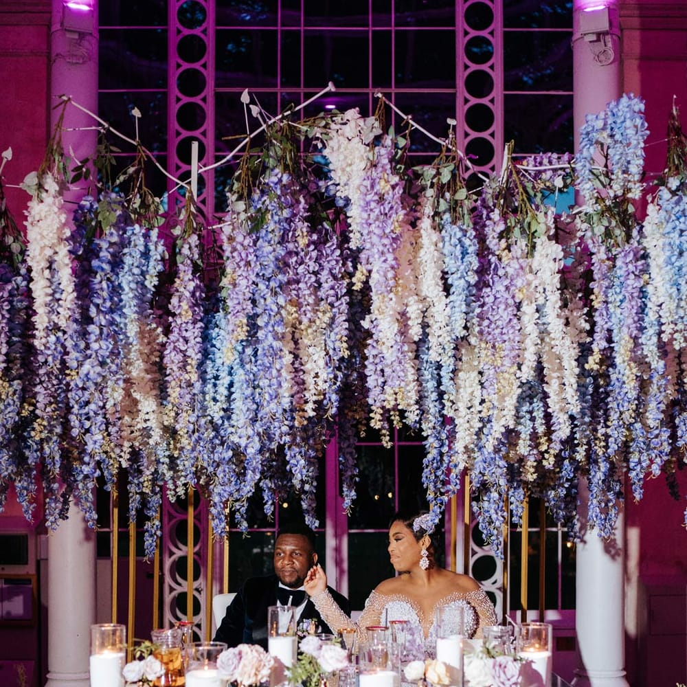 Bride in illusion v neck sheer off shoulder long sleeve pearl embellished wedding gown sitting with groom in velvet midnight blue double breasted groom’s blazer, velvet bowtie and peach garden rose boutonniere at wedding top table with suspended pastel flower decor, hurricane vases and pillar candles, gold flower vases, pastel flower centrepieces and pastel and foliage flower arrangements 