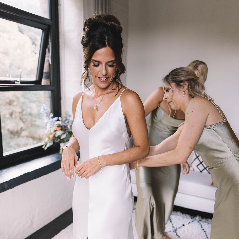 Bridesmaid in sage green satin bridesmaid dress helping bride into her satin wedding gown 