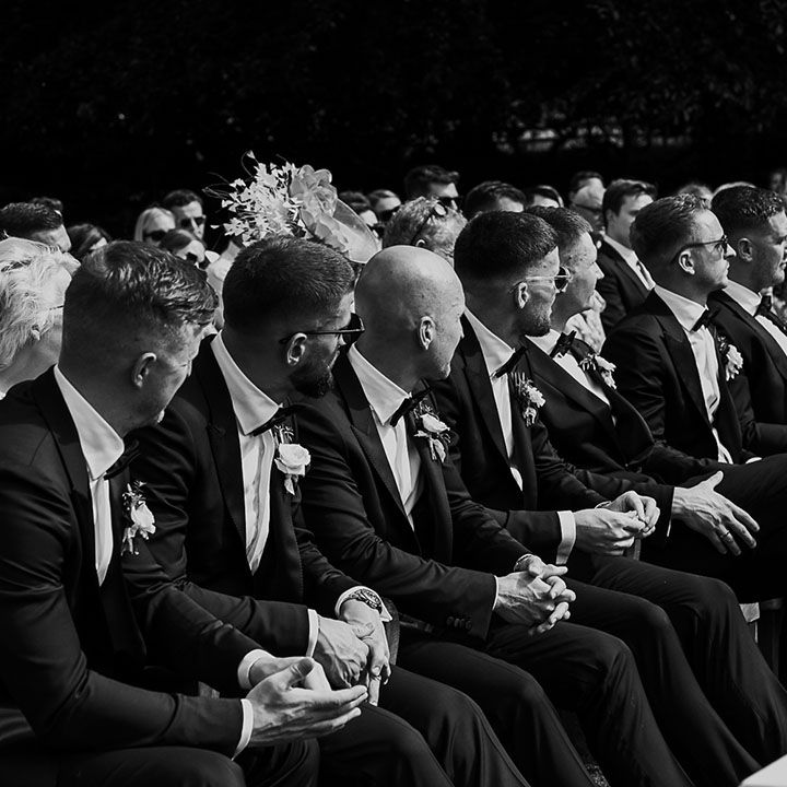Groomsmen in black tuxedos sitting together at the ceremony 