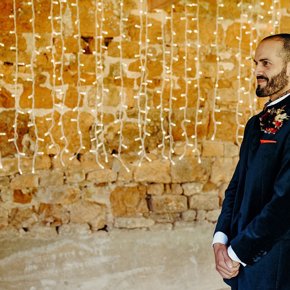 Groom in blue velvet jacket stands in front of fairy lights curtain 