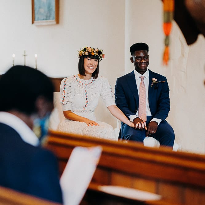 Bride in a Broderie Anglaise wedding dress resting her hand on her grooms knee during the church wedding ceremony 
