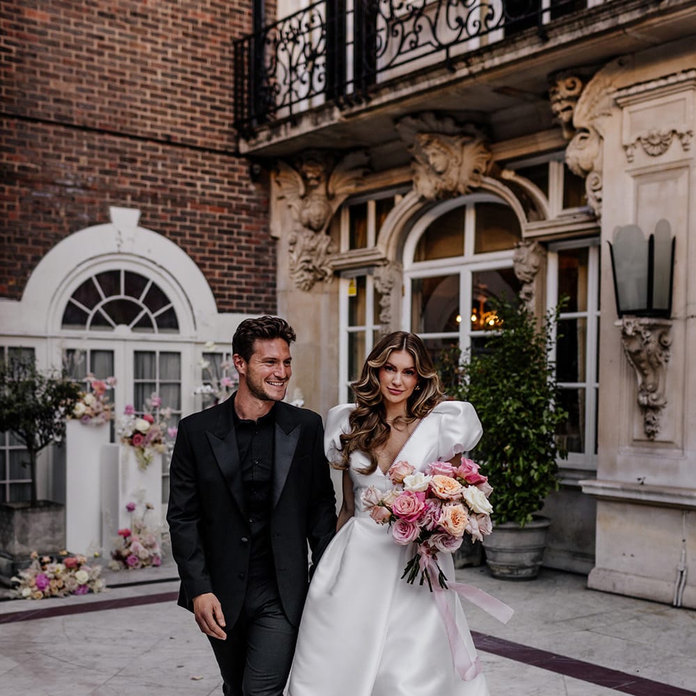 Bride in a Love in Lace bridal gown holding a pink peeled back rose wedding bouquet tied with ribbon and her husbands hand dressed in black 