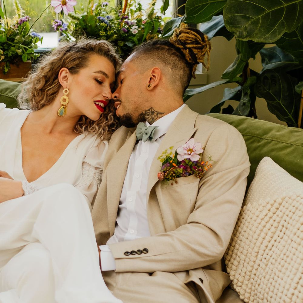 Groom in a beige suit with mint green bow tie and wildflower buttonhole flowers kissing his curly haired bride on a velvet green sofa in a bridal jumpsuit 