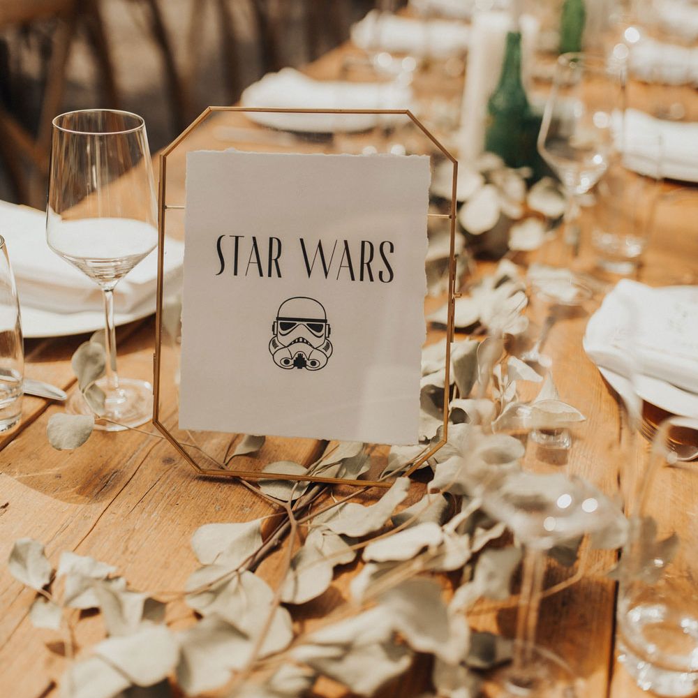 Gold framed Star Wars table name with stormtrooper helmet illustration on wooden table with foliage table garland, tableware and candles for glasshouse wedding breakfast at Anran Devon