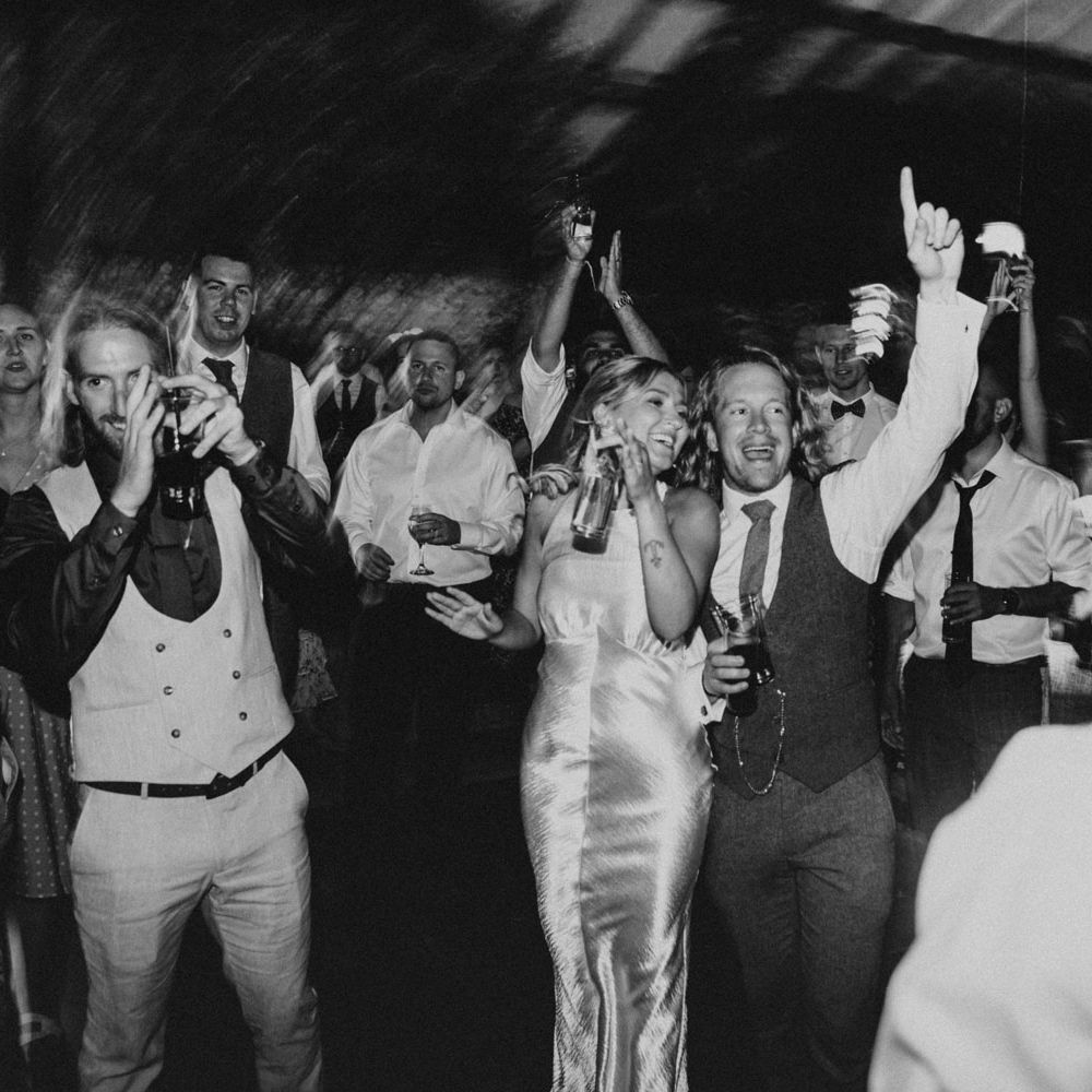 Bride in silver satin dress holding drink dances with wedding guests in the glasshouse at Anran Devon for wedding evening party