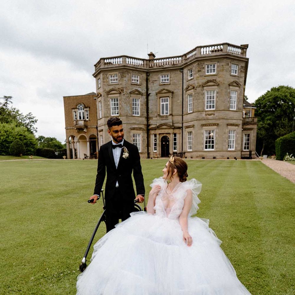Portrait of a disabled bride and groom in a wheelchair and with a walking frame outside their country house wedding venue 