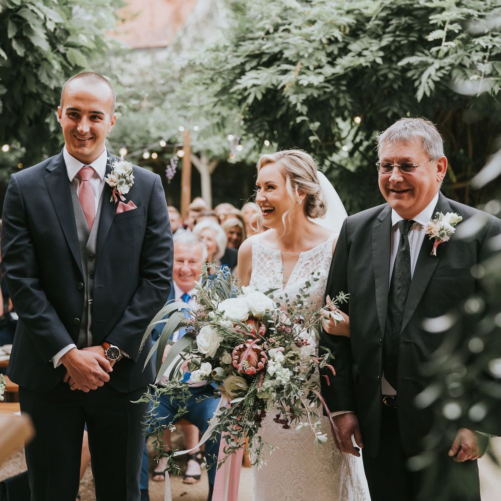 Groom in dark three piece suit with grey waistcoat and pink tie stands with hands clasped next to bride in lace Justin Alexander wedding dress holding mixed white and pink bridal bouquet arm in arm with father of the bride in black suit during outdoor wedding ceremony at Tythe Barn