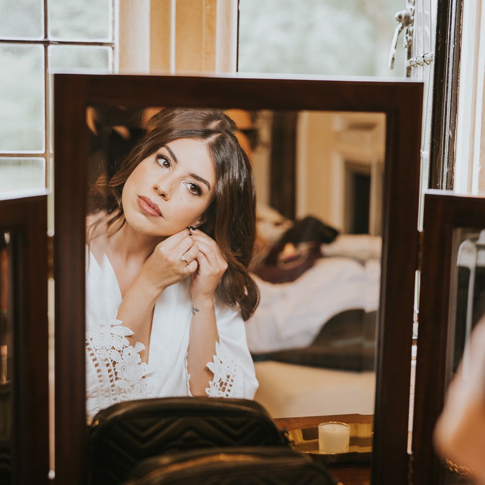 Bride in white lace trimmed bridal robe puts in her earrings as she looks in mirror at Notley Abbey wedding