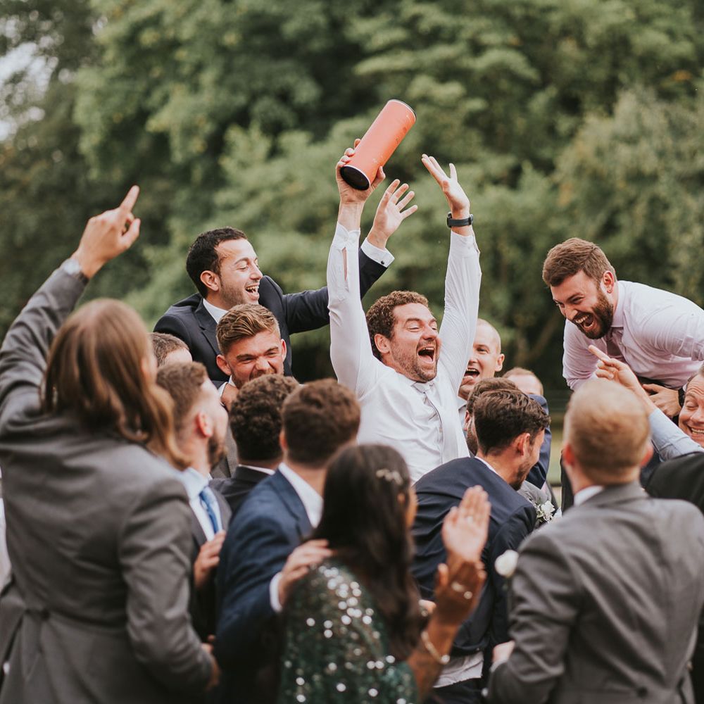 Group of wedding guests all cheer as man in white shirt and white tie catches whisky bottle box at Notley Abbey wedding