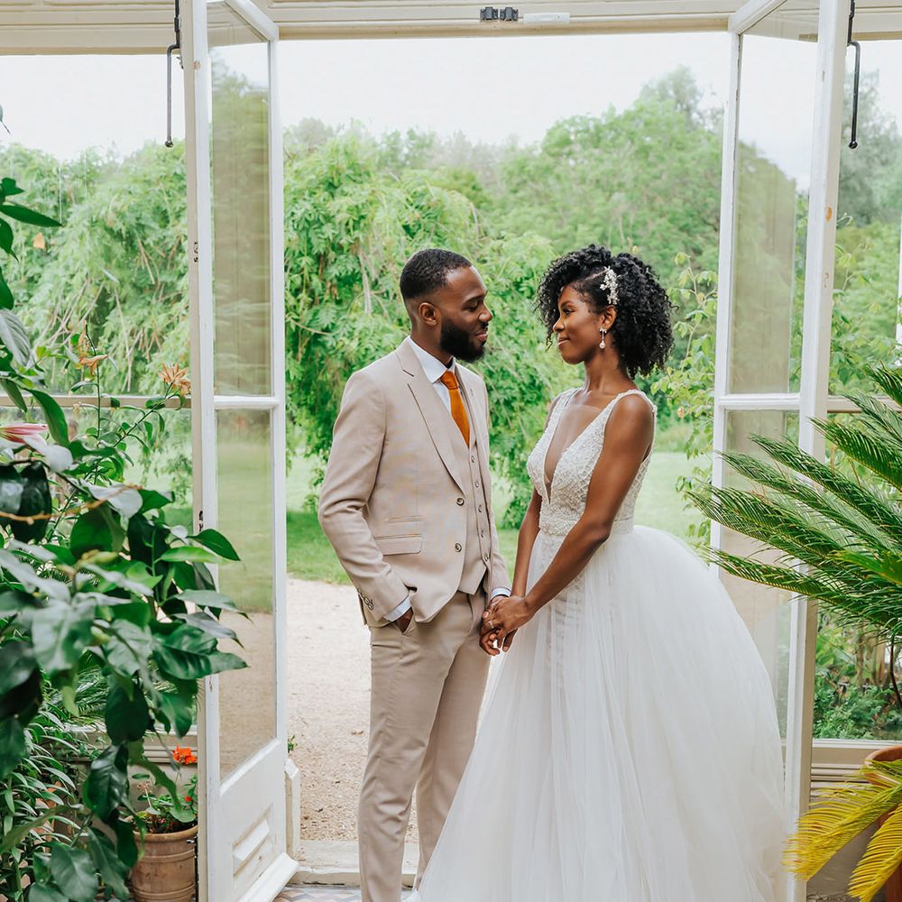 Black bride in a princess, plunging neckline wedding dress holding hands with her groom in beige suit in a conservatory at Modern Hall, London