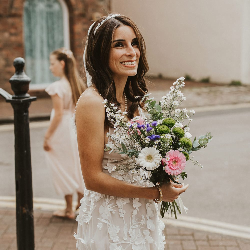 Bride smiles and holds floral bouquet and wears her brown hair down in natural curled style