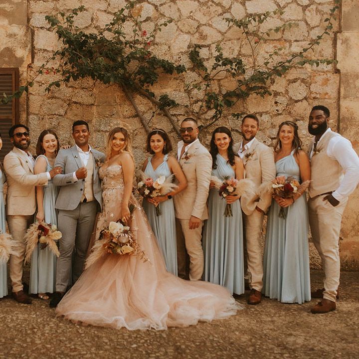 Bride & groom stand with their wedding party who wear dusty blue bridesmaid gowns and pale suits