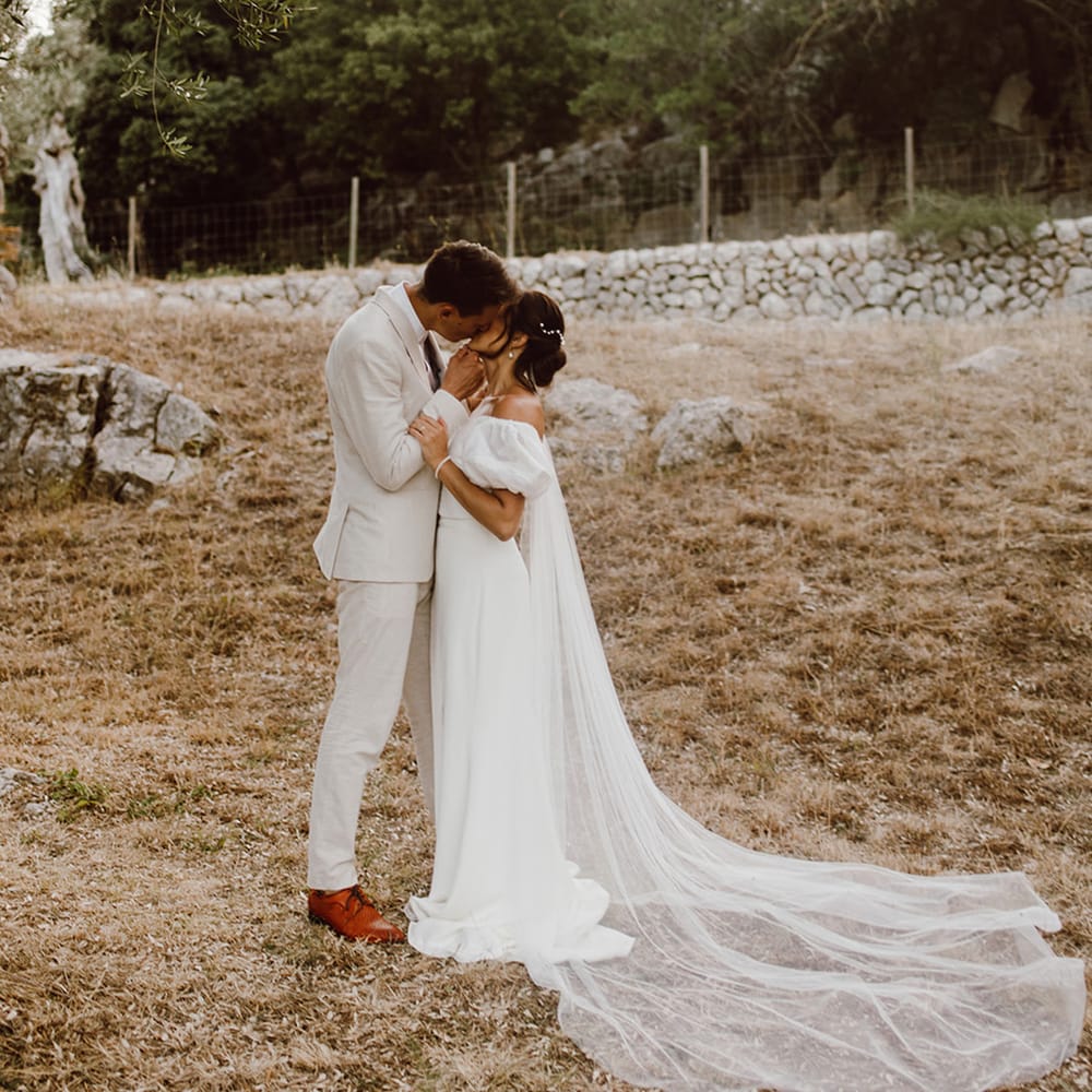 Bride in off the shoulder puff sleeve Alexandra Grecco dress shares a kiss with groom in beige suit in Mallorca