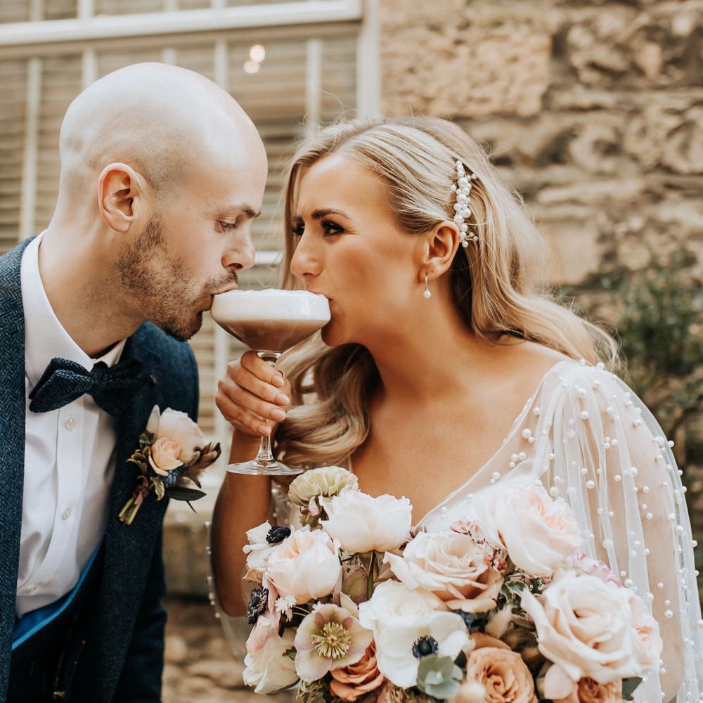 Bride with pearl detail wedding dress and pearl hair clip sits on espresso martini with groom in three piece tweed suit with bow tie