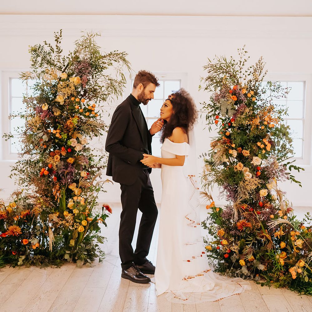 Groom in all black touching his brides face in an off the shoulder wedding dress in between orange and green column wedding ceremony flower arrangements 